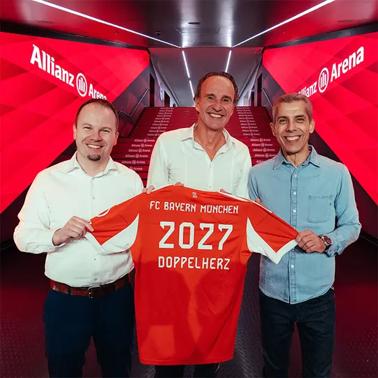 三位男士在安联竞技场前合影，展示一件印有“FC Bayern München 2027 DOPPELHERZ”的足球衣。背景为红色灯光和竞技场标志，传达出兴奋和庆祝的氛围。